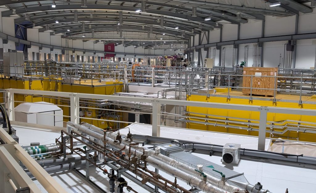 View inside the Diamond Light Source showing metal piping systems and insulated tubes in the foreground, mounted on metal supports. In the background are yellow structural elements of beamline labs and a network of walkways and railings.