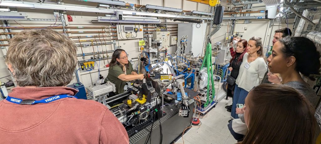 Visitors of the Harwell Heritage Network meeting observes Dr. Susan Nehzati explaining a data collection setup inside a beamline laboratory. The room is filled with scientific equipment, metal piping, cables, and control systems.